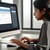 A person sitting at a desk, with a computer screen showing the HubSpot CRM dashboard on one side and the LinkedIn Sales Navigator dashboard on the other side. The person is typing on a keyboard, with a focused expression on their face.